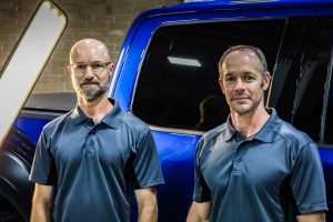 chris and coworker in polo shirts standing in front of a blue pickup truck they're working on